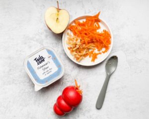 Top down shot of ingredients on a kitchen workbench: half an apple, grate carrot and apple, wet food tray, plastic spoon and filled occupier toy